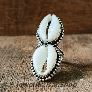 Könnte beinhalten: Ein silberner Ring mit zwei weißen Kaurischnecken. Die Muscheln sind in einen perlenbesetzten Rahmen gefasst. Der Ring wird auf einer Holzoberfläche präsentiert. Der Text "JewelArtisanshop" ist unten sichtbar.