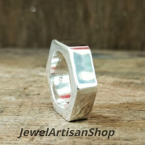 May include: A silver-colored ring with a rectangular top and a geometric design. The ring is displayed on a wooden surface. The text "JewelArtisanShop" is visible at the bottom of the image.