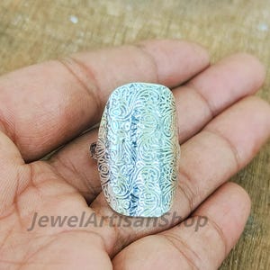 May include: A silver-colored ring with an ornate, swirling pattern. The ring is rectangular and covers a large portion of the finger. The text "JewelArtisanShop" is visible at the bottom of the image.