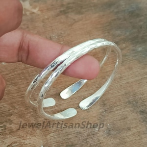 Puede incluir: Un brazalete plateado con textura martillada. El diseño abierto presenta dos bandas paralelas. El brazalete se muestra sobre una superficie de madera. El texto "JewelArtisanShop" es visible.