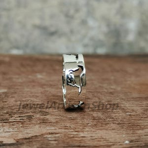 May include: A silver ring with a detailed cutout design. The ring is made of shiny metal and has a unique pattern. The ring is displayed on a wooden surface.