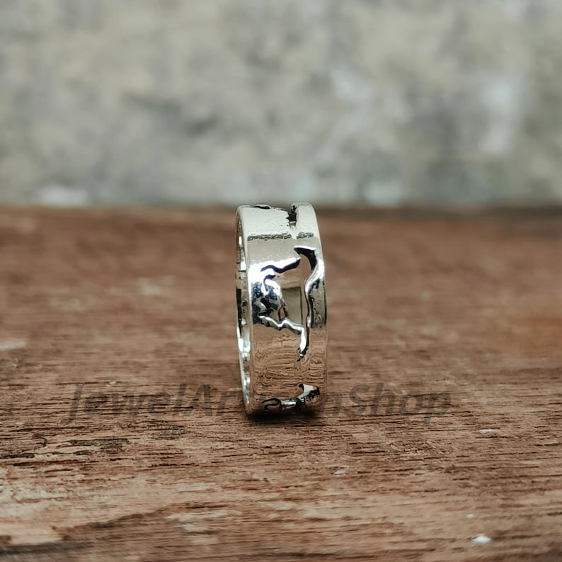May include: A silver ring with a detailed cutout design. The ring is made of shiny metal and has a unique pattern. The ring is displayed on a wooden surface.
