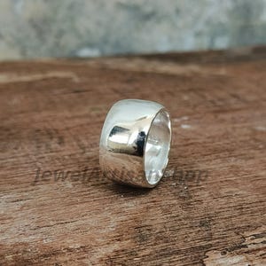 Op de afbeelding: Een brede, gepolijste zilveren ring met een glad, afgerond ontwerp. De ring reflecteert licht, waardoor een heldere, glanzende uitstraling ontstaat. De ring is een eenvoudig, maar elegant sieraad.