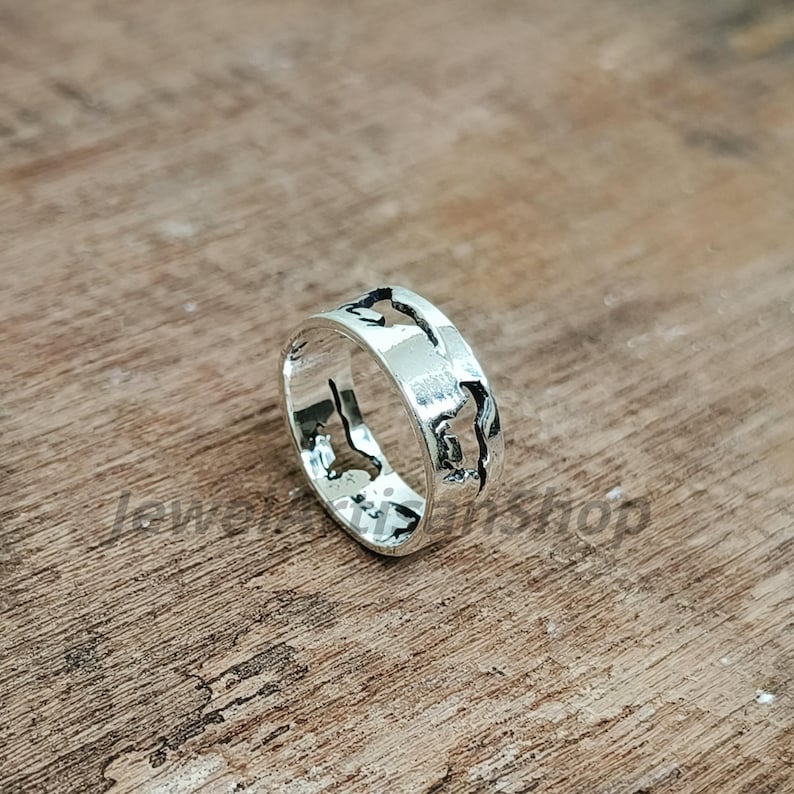 May include: A silver ring with a world map design. The ring is made of shiny metal and features cut-out shapes of continents and islands. The ring is displayed on a wooden surface.