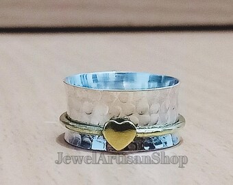 Può includere: Anello color argento con una texture martellata e un ciondolo a forma di cuore dorato. L'anello ha una fascia rotante e una fascia dorata. Il testo "JewelArtisanShop" è visibile sull'anello.