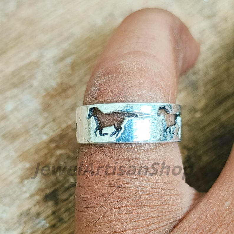 May include: A silver ring with a band featuring two brown and black horse silhouettes. The ring is worn on a finger. The text "JewelArtisanShop" is visible on the ring.