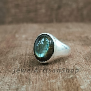 Könnte beinhalten: Ein Silberring mit einem ovalen, irisierenden Labradoritstein. Der Stein zeigt Farbtöne von Blau, Grün und Gold. Der Ring liegt auf einer Holzoberfläche. Der Text "JewelArtisanShop" ist sichtbar.