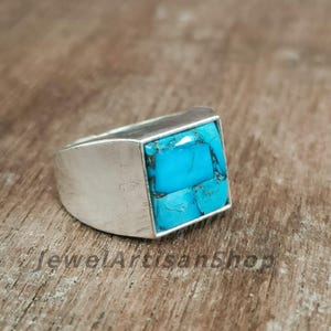 Könnte beinhalten: Ein Silberring mit einem quadratischen Türkis-Steineinsatz. Der Türkis hat eine leuchtend blaue Farbe und ist in eine silberne Lünette gefasst. Der Ring hat ein breites Band und ein modernes Design. Der Text "JewelArtisanShop" ist sichtbar.