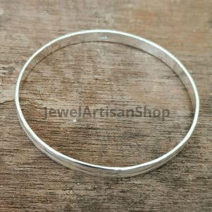 Könnte beinhalten: Ein silbernes Armreif-Armband mit glatter, polierter Oberfläche. Das Armband ist kreisförmig und scheint aus Metall zu sein. Der Text "JewelArtisanShop" ist auf der Holzoberfläche sichtbar. Dieses Schmuckstück ist für den Alltag geeignet.