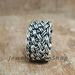 May include: A silver ring with a woven design. The ring has a thick band with an intricate pattern of interwoven metal strands. The ring is displayed on a wooden surface. The text "Jewel Shop" is visible.