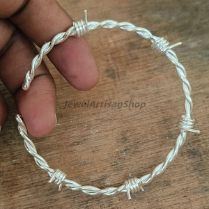 Könnte beinhalten: Ein silberfarbenes Armband, das Stacheldraht nachempfunden ist. Das Armband besteht aus verdrilltem Metall mit kleinen, spitzen Widerhaken. Das Armband wird vor einer Holzoberfläche gezeigt. Der Text "JewelArtisanShop" ist sichtbar.