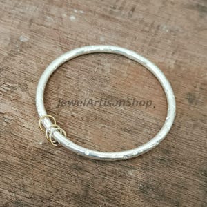 Könnte beinhalten: Ein silbernes Armreif-Armband mit strukturierter Oberfläche und einer kleinen Ansammlung goldfarbener Ringe. Das Armband ist kreisförmig und liegt auf einer Holzoberfläche. Der Text "JewelArtisanShop" ist sichtbar.