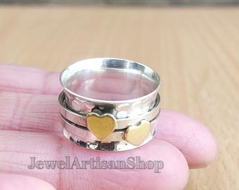 Può includere: Un anello color argento con due cuori dorati. L'anello ha una fascia strutturata e una parte superiore girevole. Il testo "JewelArtisanShop" è visibile nella parte inferiore dell'immagine.