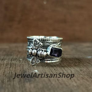 Könnte beinhalten: Silberfarbener Ring mit einer Biene und einem rechteckigen, lila Edelstein. Der Ring weist aufwendige Details auf, und der Text "JewelArtisanShop" ist sichtbar. Der Ring liegt auf einer Holzoberfläche.