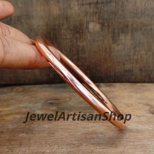 Könnte beinhalten: Zwei polierte Kupferarmreifen werden gezeigt, die ihre glatte, reflektierende Oberfläche zeigen. Die Armreifen haben eine warme, rötlich-braune Farbe, und der Text "JewelArtisanShop" ist unten sichtbar.