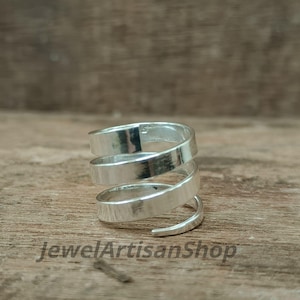 Puede incluir: Un anillo en espiral plateado con un diseño de banda plana. El anillo se envuelve alrededor del dedo varias veces, creando un efecto en capas. El anillo se muestra sobre una superficie de madera. El texto "JewelArtisanShop" es visible.
