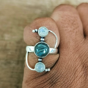 Könnte beinhalten: Ein silberner Ring mit drei Edelsteinfassungen. Die mittlere Fassung hält einen runden, teal-farbenen Stein, flankiert von zwei kleineren, runden, klaren Steinen. Der Ring hat ein einzigartiges, offenes Design mit geschwungenen silbernen Akzenten.