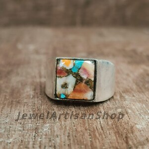 Könnte beinhalten: Ein Silberring mit einem quadratischen, eingelegten Stein. Der Stein weist ein Mosaik aus Farben auf, darunter Weiß, Orange, Türkis und Braun. Der Ring hat ein breites Band und eine polierte Oberfläche. Der Text "JewelArtisanShop" ist sichtbar.
