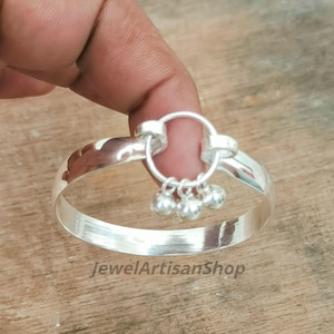 May include: A silver bangle bracelet with a small, dangling charm featuring three tiny bells. The bracelet is held by a hand, showcasing its polished, reflective surface. The text "JewelArtisanShop" is visible.