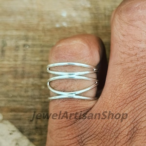 Könnte beinhalten: Ein silberfarbener Ring mit offenem, geschichtetem Design. Der Ring hat mehrere Bänder, die sich kreuzen und einen komplizierten und modernen Look erzeugen. Der Ring wird an einem Finger gezeigt, um seine Form und seinen Stil hervorzuheben. Der Ring ist ein Schmuckstück.