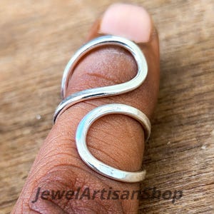 Puede incluir: Un anillo plateado con un diseño único y fluido. El anillo se envuelve alrededor de un dedo, con una parte superior curvada y una banda inferior circular. El anillo está hecho de metal brillante y se muestra en el dedo de una persona. El texto "JewelArtisanShop" es visible.
