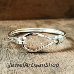 May include: A silver-colored bracelet with a hook-and-eye clasp. The bracelet is made of thin, round metal and features a looped design. The text "JewelArtisanShop" is visible at the bottom of the image.