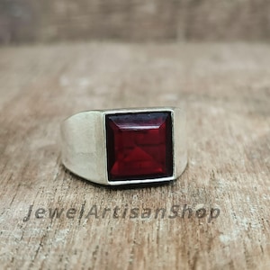 Könnte beinhalten: Ein silberner Ring mit einem quadratischen, tiefroten Edelstein. Der Ring hat ein breites Band und eine Lünettenfassung für den Stein. Der Text "JewelArtisanShop" ist auf der Holzoberfläche sichtbar. Dieser Ring ist ein Schmuckstück.