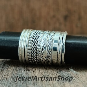 Może przedstawiać: Kolekcja srebrnych pierścionków o różnych wzorach, w tym plecionych, koralikowych i kwiatowych. Pierścionki są prezentowane na czarnym cylindrycznym obiekcie. Tekst "JewelArtisanShop" jest widoczny na dole.