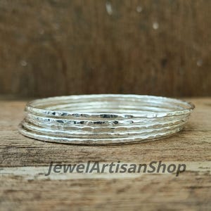 Könnte beinhalten: Ein Stapel silberner Armreifen mit gehämmerten Texturen, auf einer Holzoberfläche. Die Armreifen sind dünn und haben ein glänzendes Finish. Der Text "JewelArtisanShop" ist unten sichtbar.
