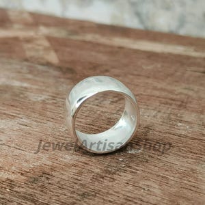Op de afbeelding: Een brede, zilveren ring met een gehamerde textuur. De ring is gemaakt van een glanzend, reflecterend metaal en heeft een gladde binnenkant. De ring is een eenvoudig, maar elegant sieraad.