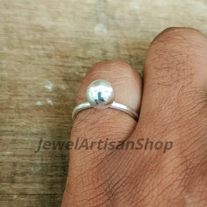 Puede incluir: Anillo de plata con una perla esférica pulida como pieza central. El anillo tiene una banda delgada y simple, y la perla refleja la luz. El texto "JewelArtisanShop" es visible en la imagen.