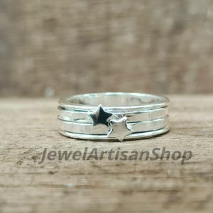 Könnte beinhalten: Ein silberner Ring mit Sternmuster. Der Ring hat zwei Sterne und drei Bänder. Der Ring liegt auf einer Holzoberfläche. Der Text "JewelArtisanShop" ist auf der Oberfläche zu sehen.