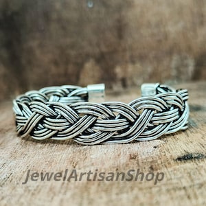 Könnte beinhalten: Ein silberfarbenes Armband mit geflochtenem Design. Das Armband hat ineinander verwobene Stränge, die ein geflochtenes Aussehen erzeugen. Der Verschluss ist oben sichtbar. Der Text "JewelArtisanShop" befindet sich unten.