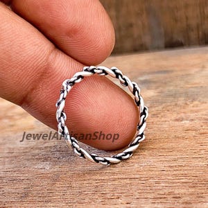 Könnte beinhalten: Ein silberfarbener Ring mit geflochtenem Seildesign. Der Ring ist kreisförmig und scheint aus Metall zu sein. Der Ring wird von den Fingern einer Person gehalten. Der Text "JewelArtisanShop" ist auf dem Bild zu sehen.