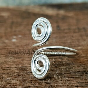 Könnte beinhalten: Ein silberfarbener Ring mit einem einzigartigen Spiraldesign. Der Ring hat zwei wirbelnde, offene Spiralen, die durch ein glattes Band verbunden sind. Der Ring ist vor einem Holzgrund platziert.