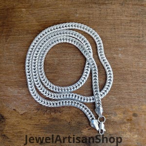 May include: A silver-colored chain necklace with a woven design, lying on a wooden surface. The necklace is coiled and features a clasp closure. The text "JewelArtisanShop" is visible at the bottom of the image.