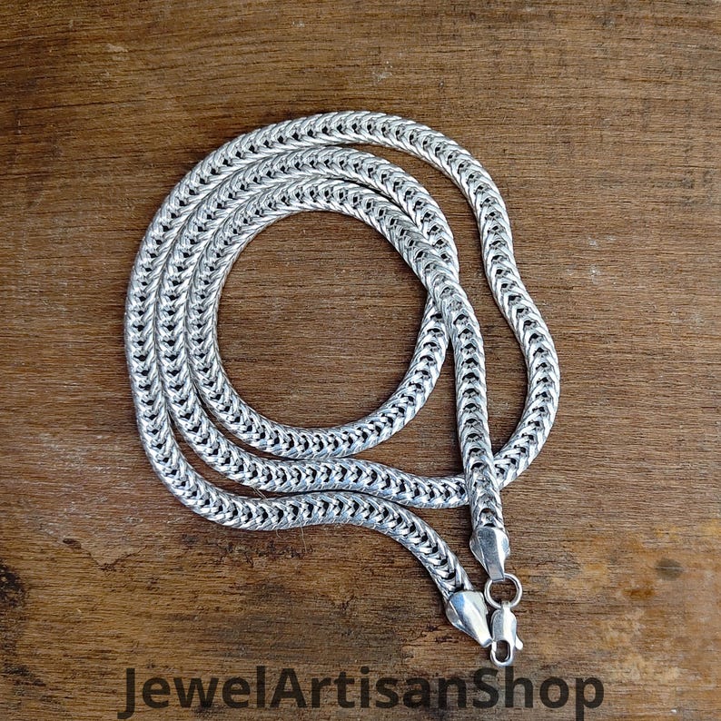 May include: A silver-colored chain necklace with a woven design, lying on a wooden surface. The necklace is coiled and features a clasp closure. The text "JewelArtisanShop" is visible at the bottom of the image.