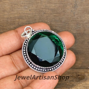 May include: A round, green gemstone pendant with a silver bezel. The faceted gemstone reflects light. The pendant has a loop for a chain. The text "JewelArtisanShop" is visible at the bottom of the image.
