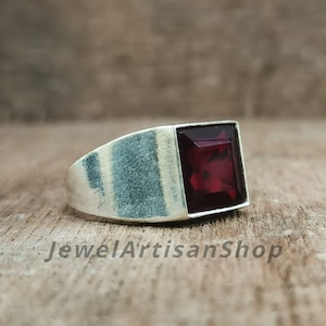 May include: A silver ring with a large, square, deep red gemstone. The ring has a wide band and a simple, modern design. The text "JewelArtisanShop" is visible on the surface.