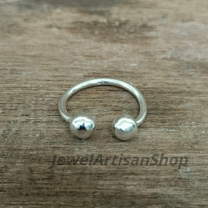 Könnte beinhalten: Ein silberner Ring mit einem gebogenen Band und zwei kugelförmigen Enden. Der Ring liegt auf einer Holzoberfläche. Der Text "JewelArtisanShop" ist unten im Bild zu sehen.