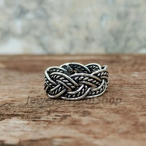 May include: A silver-colored ring with a braided design. The ring features an intricate pattern of interwoven strands, creating a textured and detailed appearance. The ring is displayed on a wooden surface.
