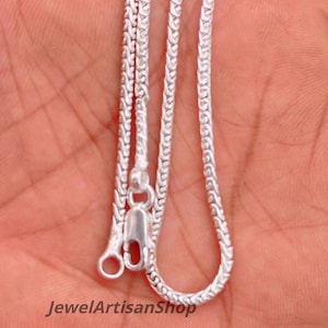 May include: A silver-colored wheat chain necklace featuring a polished finish and a lobster claw clasp. The chain consists of interconnected, braided metal links that create a textured, rope-like appearance. The necklace includes a small circular jump ring.