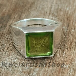 Könnte beinhalten: Ein silberner Ring mit einem quadratischen, hellgrünen Edelstein. Der Ring hat ein dickes Band und eine polierte Oberfläche. Der Edelstein ist in einer quadratischen Lünette gefasst und hat ein leicht strukturiertes Aussehen. Der Ring wird auf einer Holzoberfläche präsentiert.