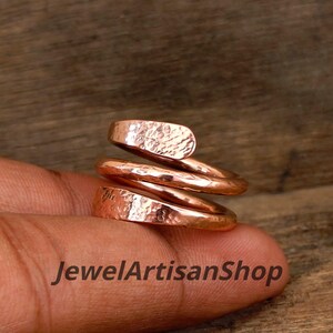 Könnte beinhalten: Ein kupferfarbener Spiralring mit gehämmerten Textur. Der Ring wickelt sich in einem einzigartigen Design um den Finger. Der Text "JewelArtisanShop" ist unten im Bild zu sehen.