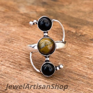 Könnte beinhalten: Ein silberner Ring mit drei kreisförmigen Steinen. Die oberen und unteren Steine sind schwarz, während der mittlere Stein eine Mischung aus Gelb und Braun aufweist. Der Ring hat ein einzigartiges, offenes Design mit gebogenen silbernen Akzenten. Der Text "JewelArtisanShop" ist sichtbar.