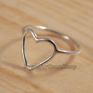 May include: A silver ring with a heart-shaped design. The ring features a delicate, open heart shape, crafted from a shiny, metallic material. The ring is set against a light brown wooden surface. The text "JewelArtisanShop" is visible on the ring.