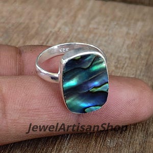 Puede incluir: Un anillo de plata con una piedra rectangular de concha de abulón. La concha iridiscente muestra tonos de verde, azul y negro. El anillo está en un dedo, con el texto "JewelArtisanShop" visible.