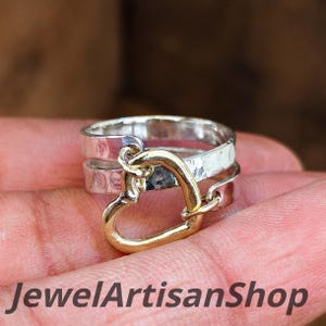 Könnte beinhalten: Ein silberner Ring mit einem goldenen Herzanhänger. Der Ring hat eine gehämmerten Textur und einen goldenen Herzanhänger. Der Text "JewelArtisanShop" ist unten im Bild zu sehen.