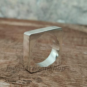 Könnte beinhalten: Ein silberfarbener, quadratischer Ring mit hohler Mitte. Der Ring hat ein modernes, geometrisches Design und wird auf einer Holzoberfläche präsentiert. Die Form des Rings ist ein kühnes, minimalistisches Statement-Stück.