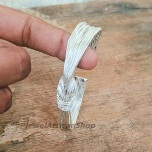 Könnte beinhalten: Ein silberfarbener Ring mit einem einzigartigen Design. Der Ring hat ein knotenartiges Detail und ein Band aus mehreren Metallsträngen. Der Ring wird vor einer Holzoberfläche gehalten.